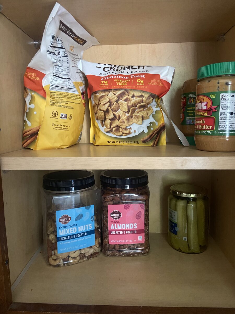 A kitchen cabinet: protein cereal, peanut butter, mixed nuts, almonds, a jar of pickles.