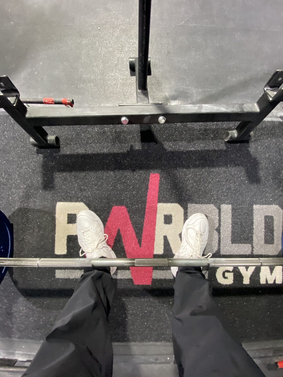 Looking straight down at a barbell on the gym floor, white sneakers either side of it, the gym's name on the mat.