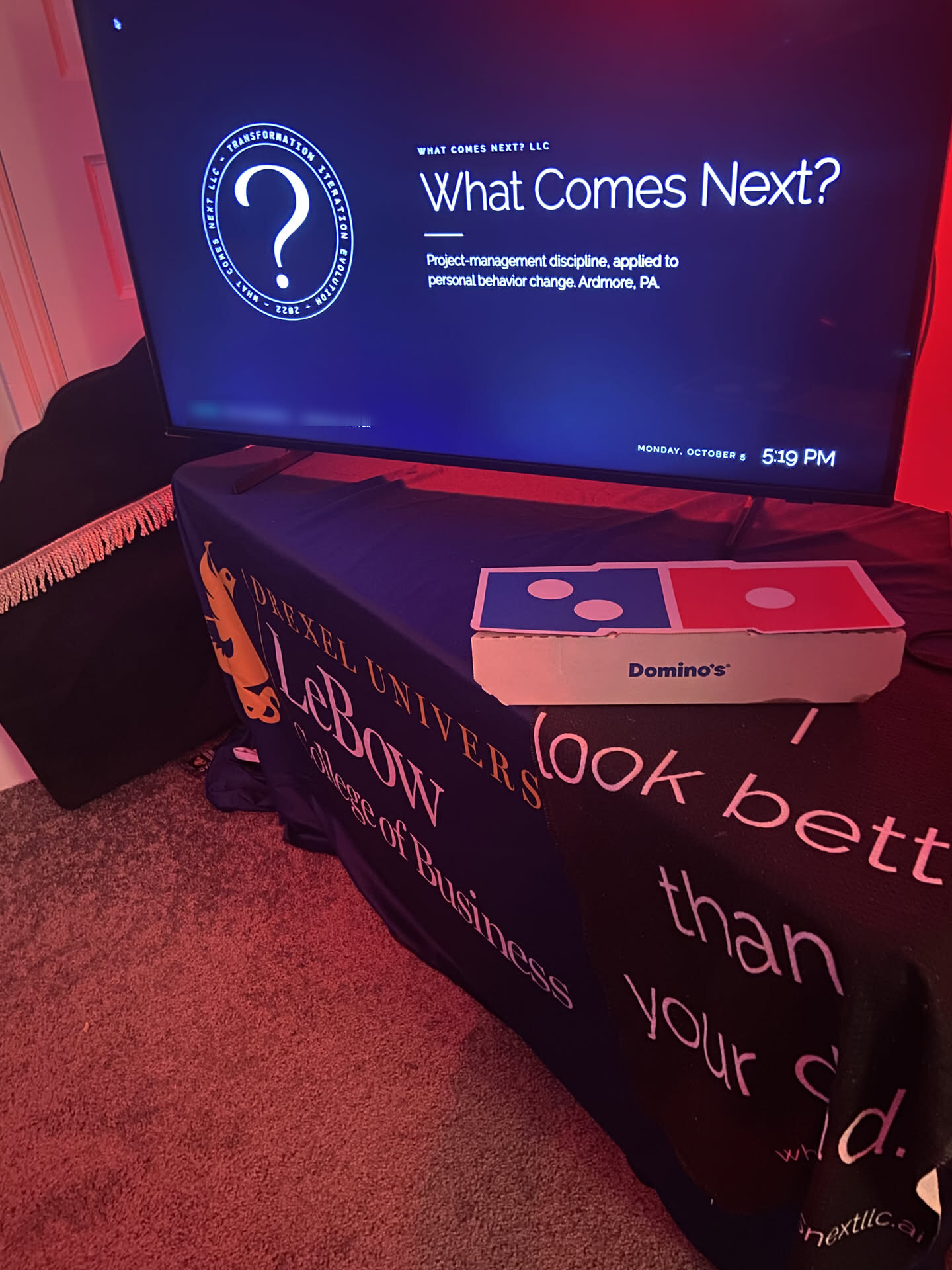 The office TV showing What Comes Next?, a Domino's box on a table draped with a Drexel LeBow banner and an I look better than your dad banner.