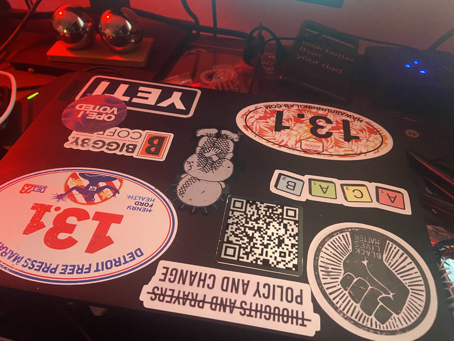 A sticker-covered laptop lid under red light: a YETI sticker, two 13.1 half-marathon stickers, a QR code, and Policy and Change.