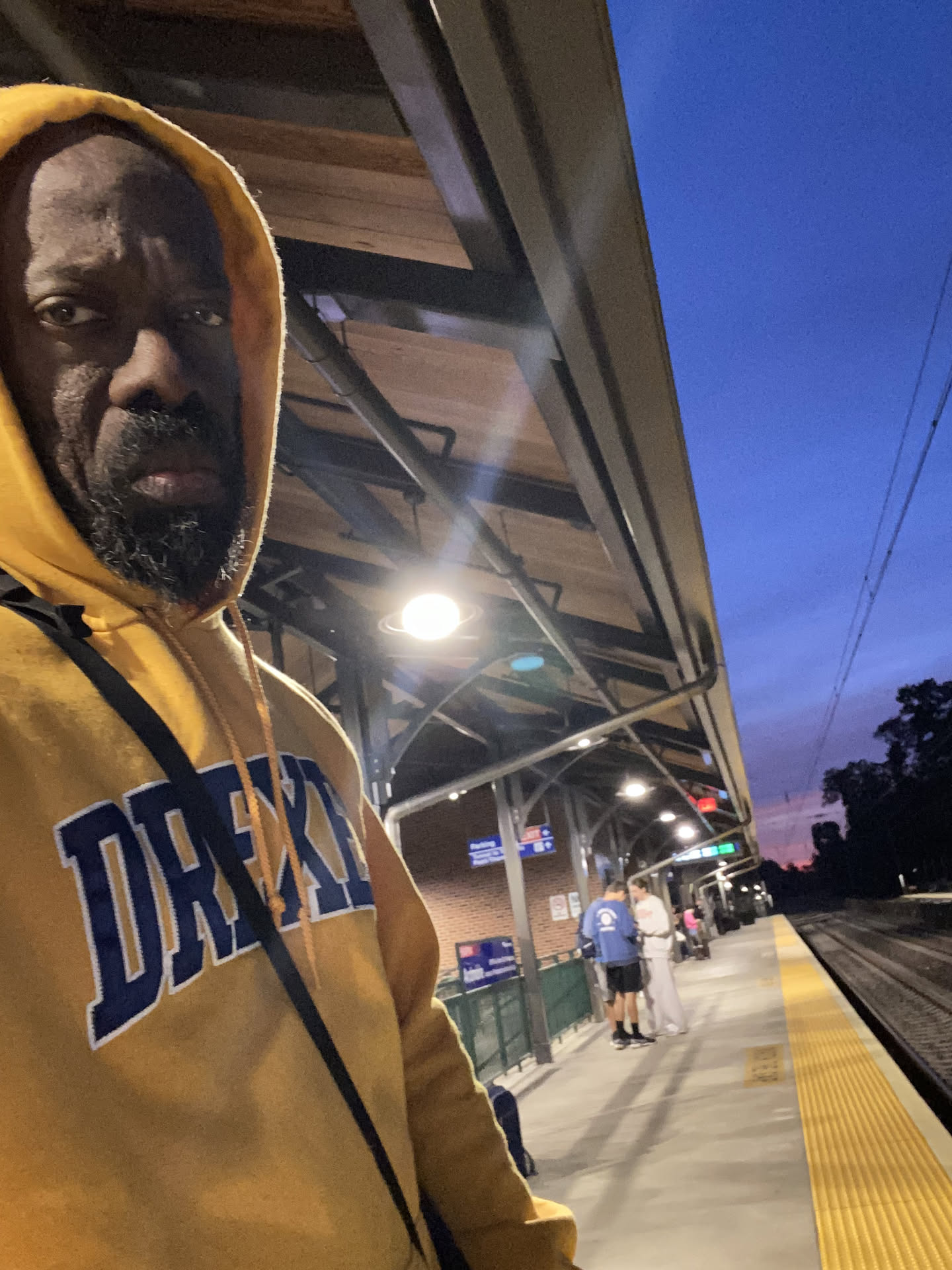 A selfie in a gold Drexel hoodie, hood up, on a train platform at dusk.