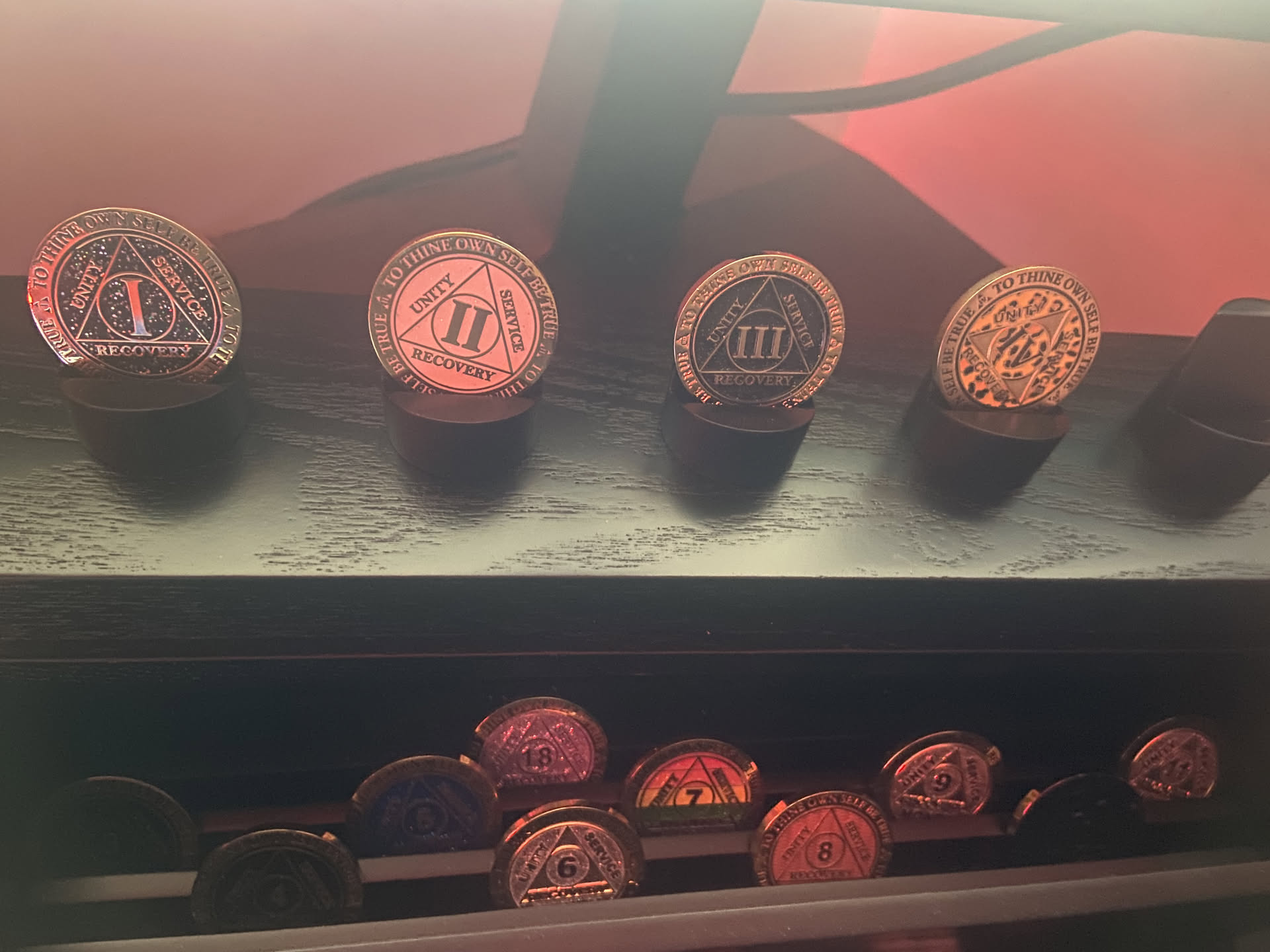 A shelf under red light: four recovery medallions on stands, I, II, III and IV; below them, a row of monthly chips.