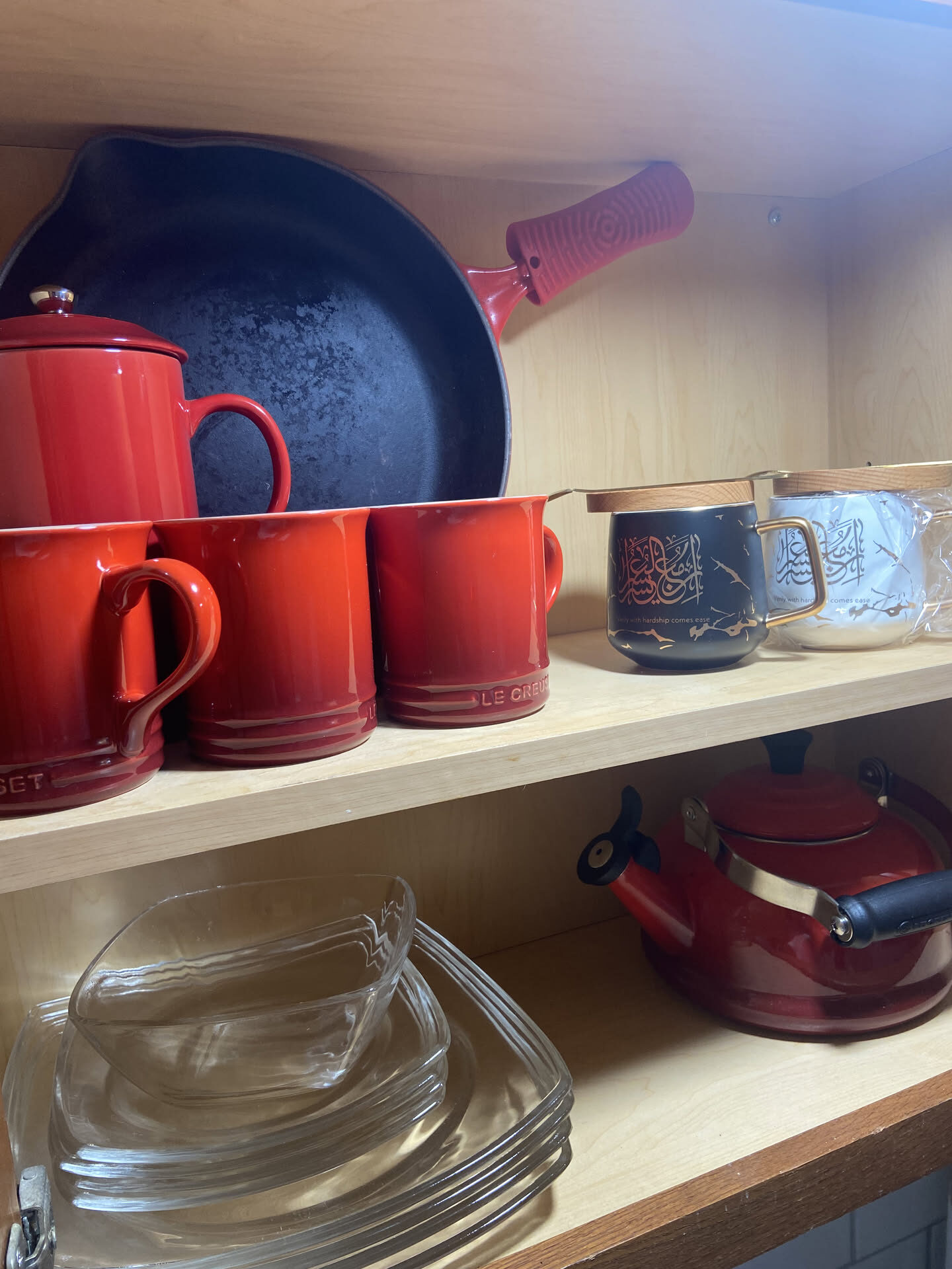An open kitchen cupboard: a red cast-iron skillet, a red coffee pot and three red mugs, two mugs with Arabic calligraphy, a red kettle, and a stack of glass plates.