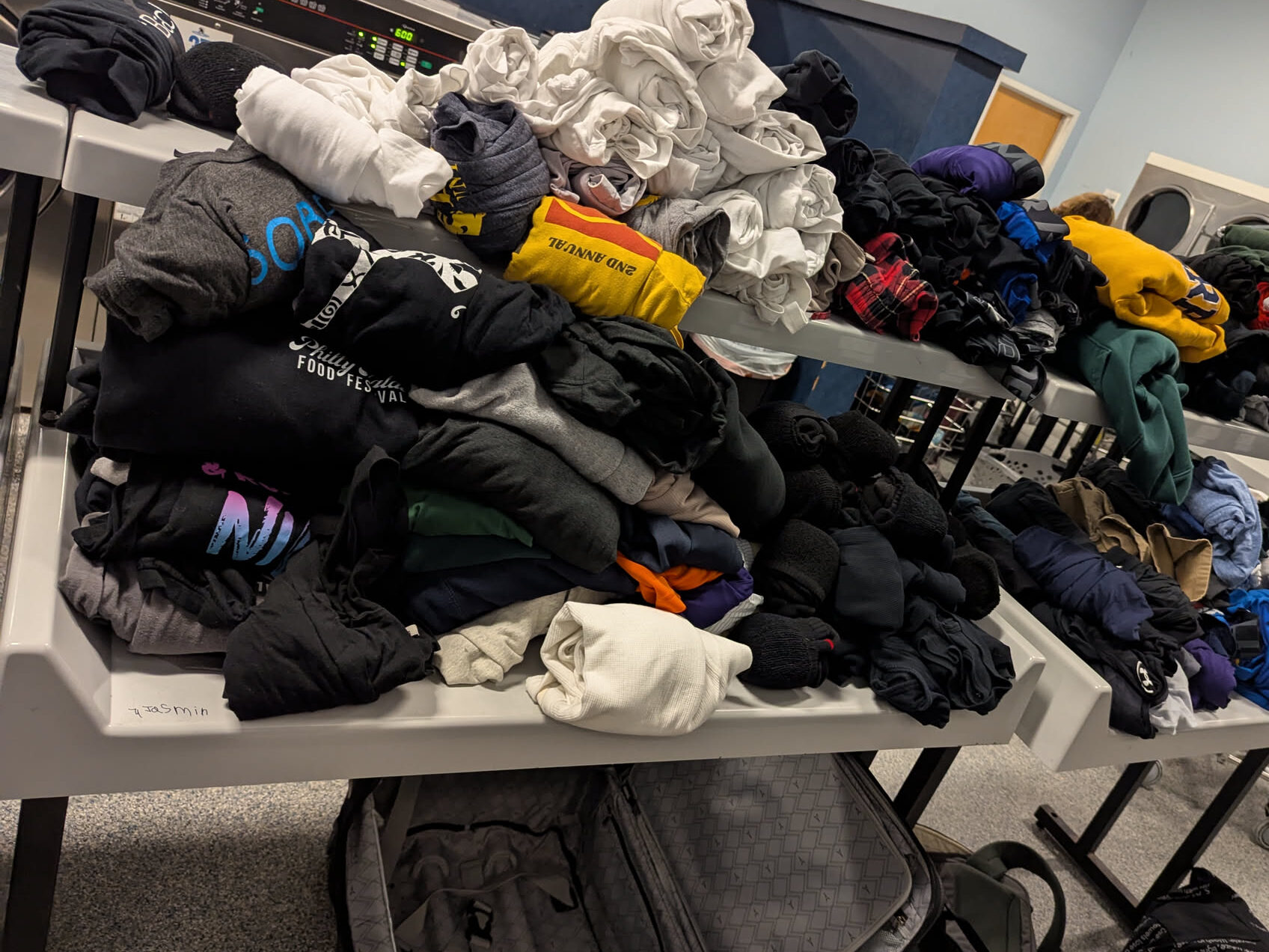 Laundromat folding tables covered end to end in folded hoodies, T-shirts and towels, an open suitcase underneath.