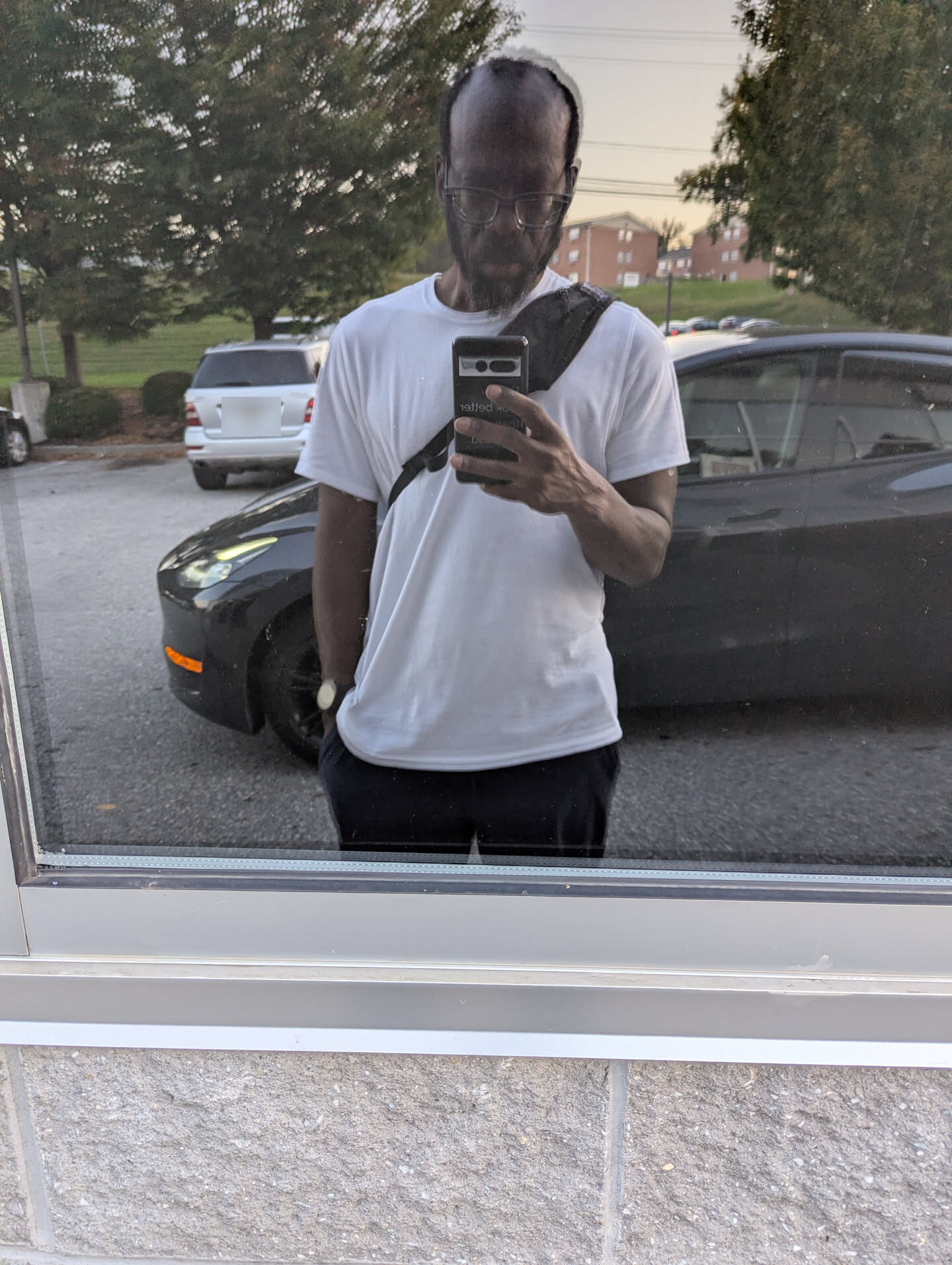 A mirror selfie in a window: white T-shirt, glasses, a sling bag across the chest, a parking lot behind.