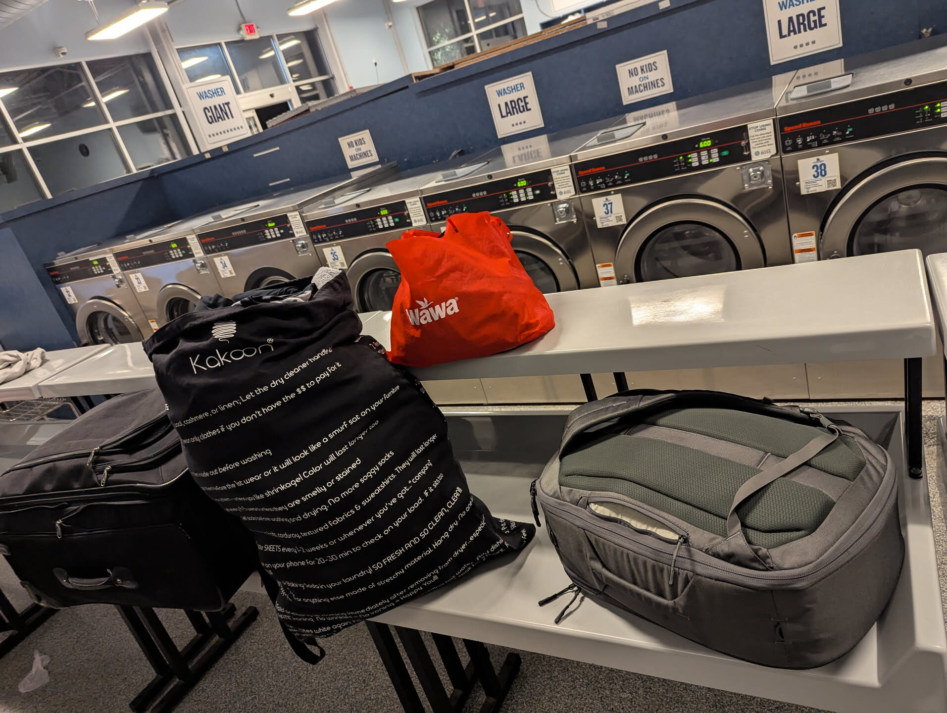 Packed up on the folding table in front of the big washers: a full laundry bag, a Wawa bag, a rolling suitcase and a backpack.