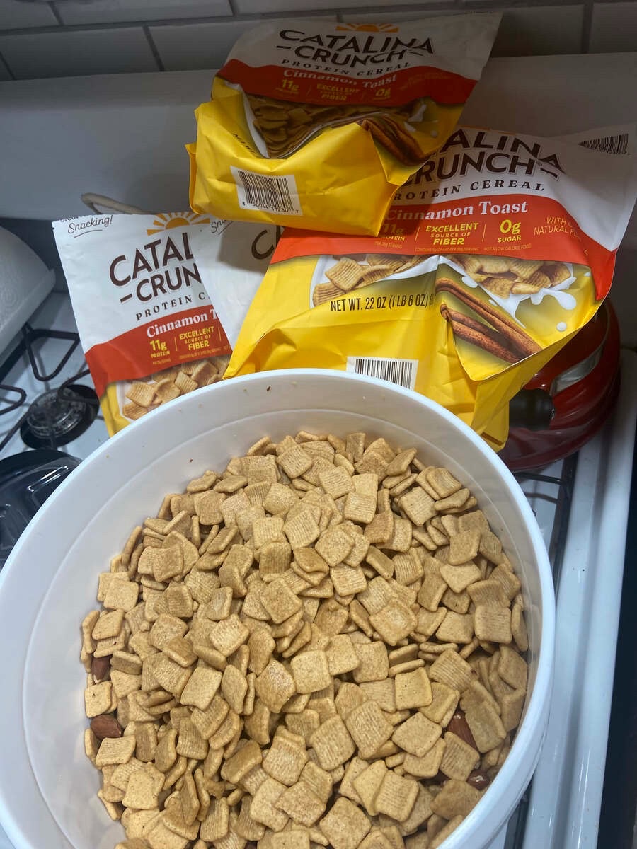 A white bucket full of protein cereal squares, lid off, with empty Catalina Crunch bags behind it: cinnamon toast.